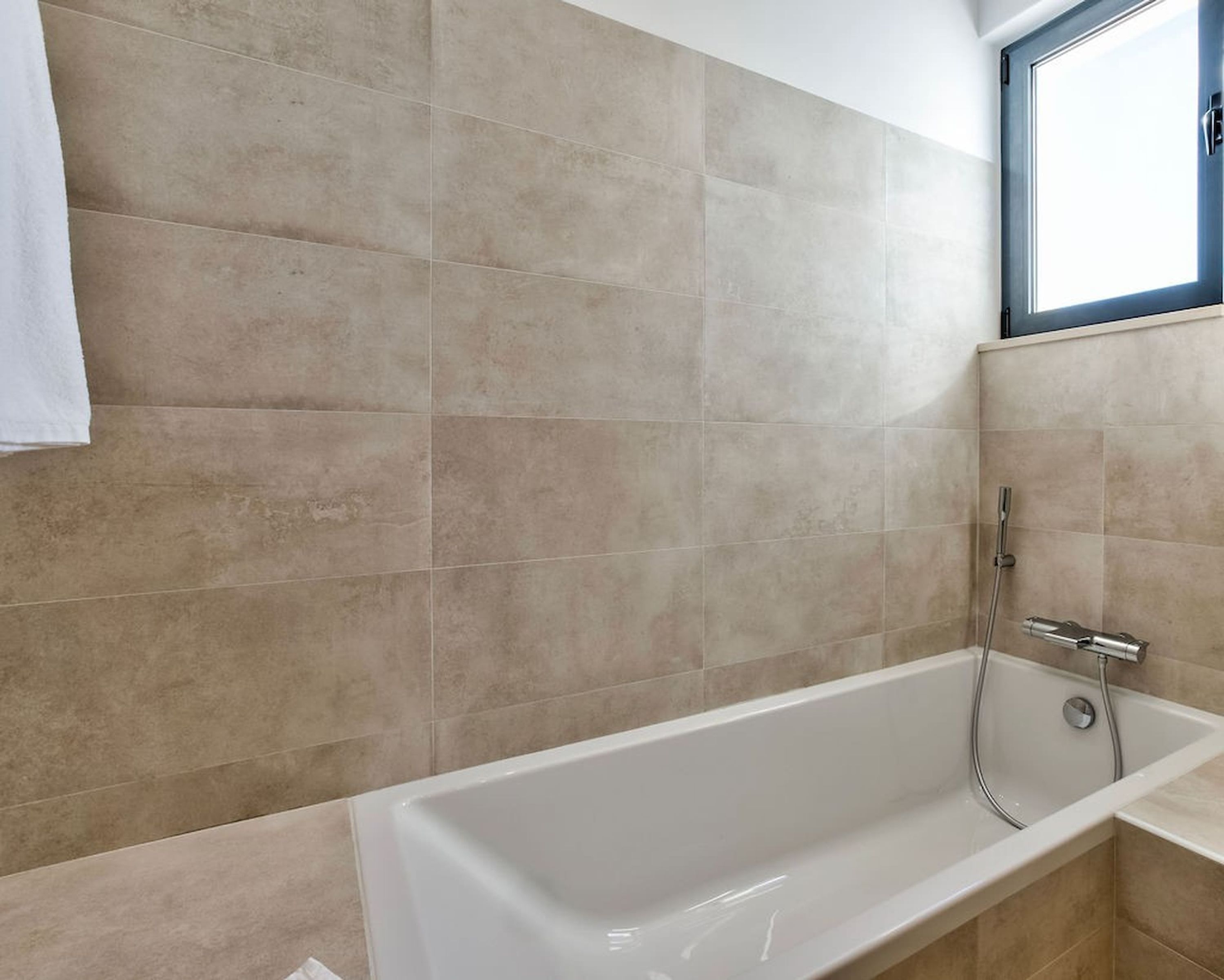 A bathroom with beige tiles, a white bathtub, and a window above the tub.