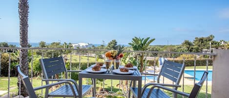 A terrace with a table set for two, overlooking a swimming pool and a lush garden.