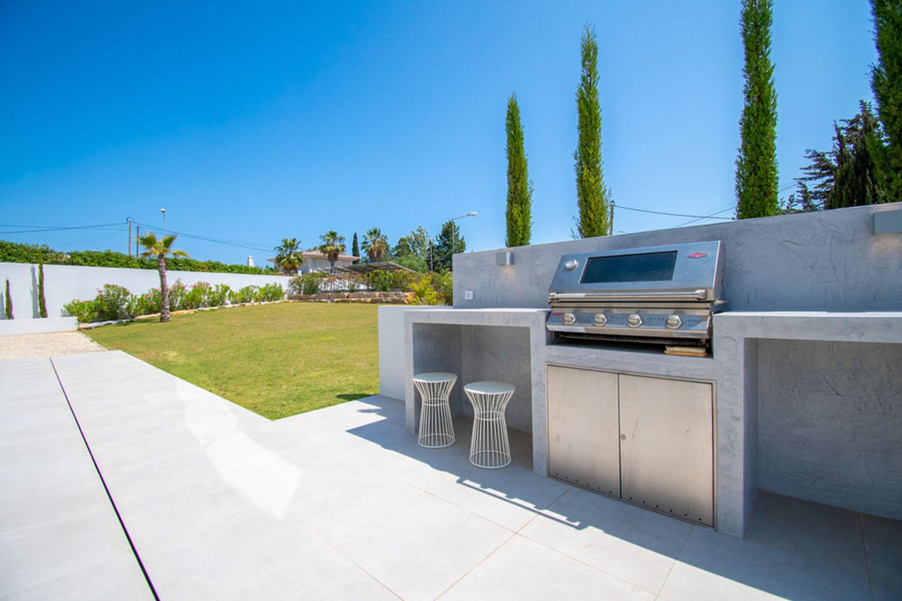 Une cuisine extérieure dotée d’un barbecue intégré et de deux tabourets, le tout installé devant un gazon bien entretenu et de grands arbres.