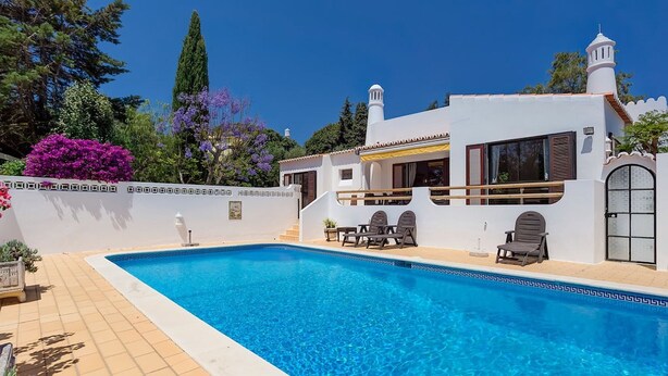 Zona de piscina con una pileta azul cristalina, un edificio blanco con balcón y un pequeño jardín con flores moradas.