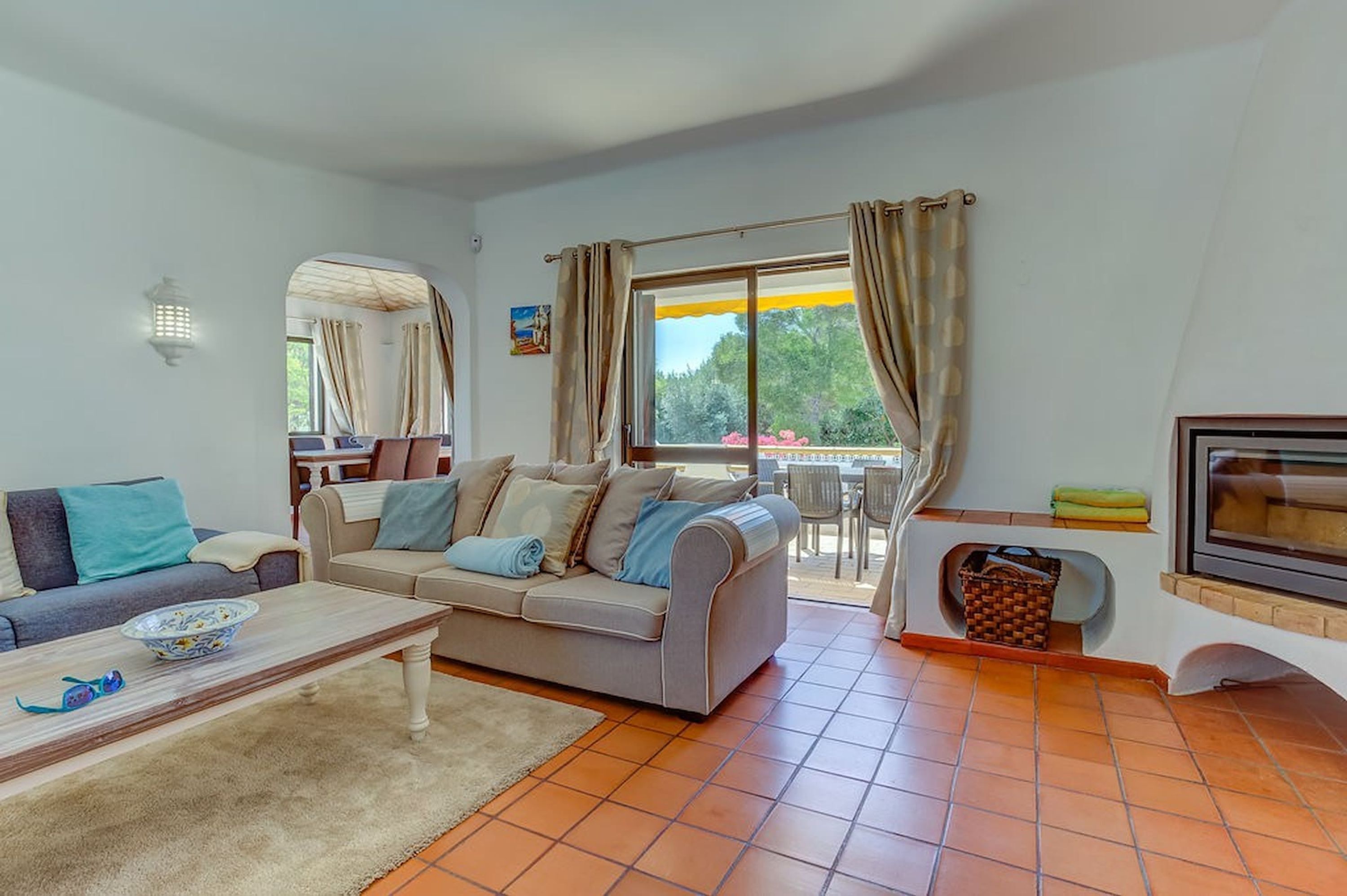 A living room with a sofa, coffee table, and a TV. There is a window with curtains, a lamp, and a view of the outdoors.