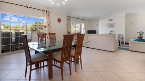 Een eetgedeelte met een glazen tafel en houten stoelen, een keuken met een wit aanrecht en een groot raam met uitzicht op groen.