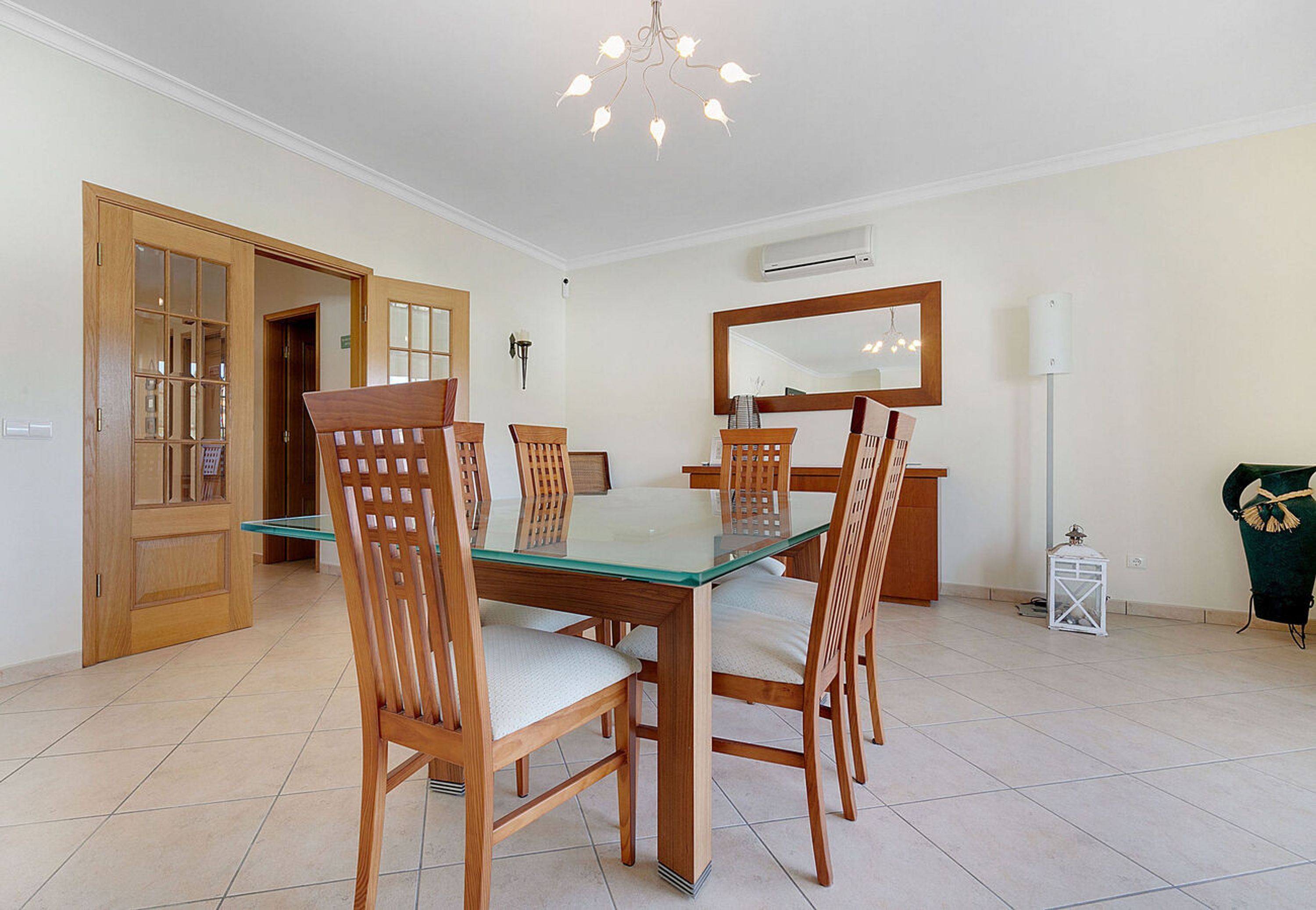 A dining area with a glass-top table, wooden chairs, a chandelier, and a large mirror on the wall.