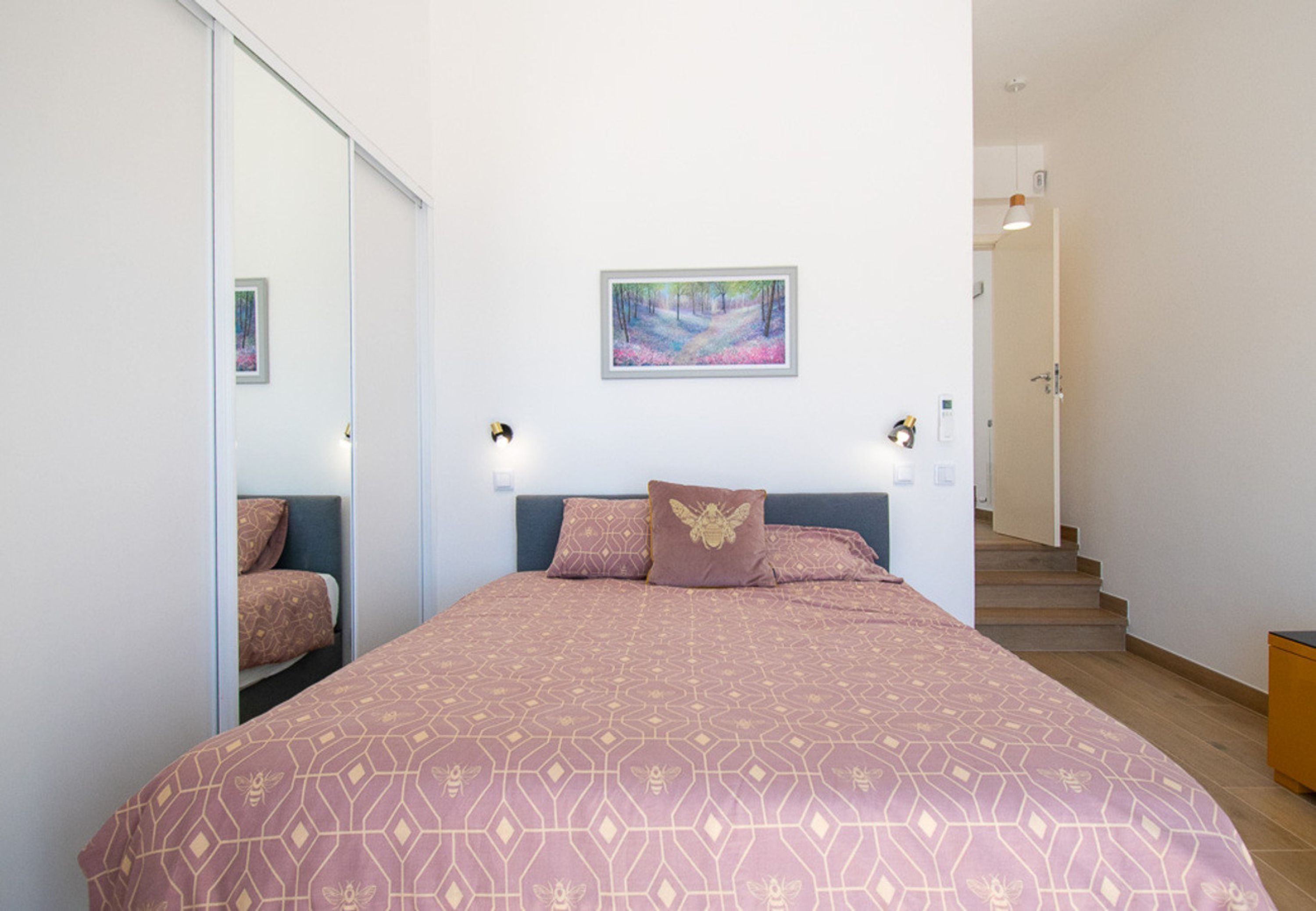 A bedroom with a large bed, a painting on the wall, and a staircase leading to another room.