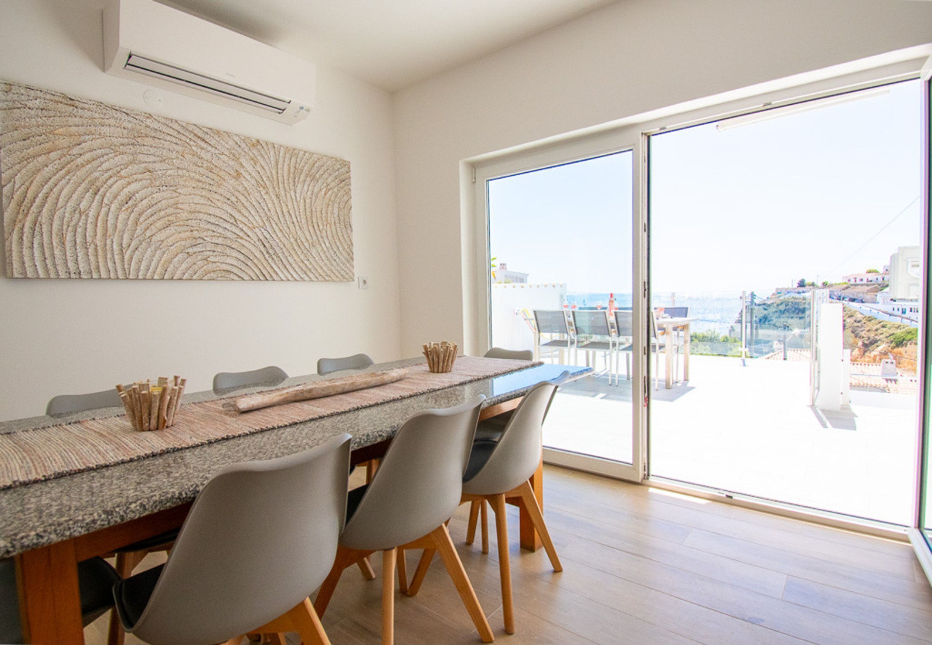 A dining area with a long table, chairs, and a view through a large glass door.