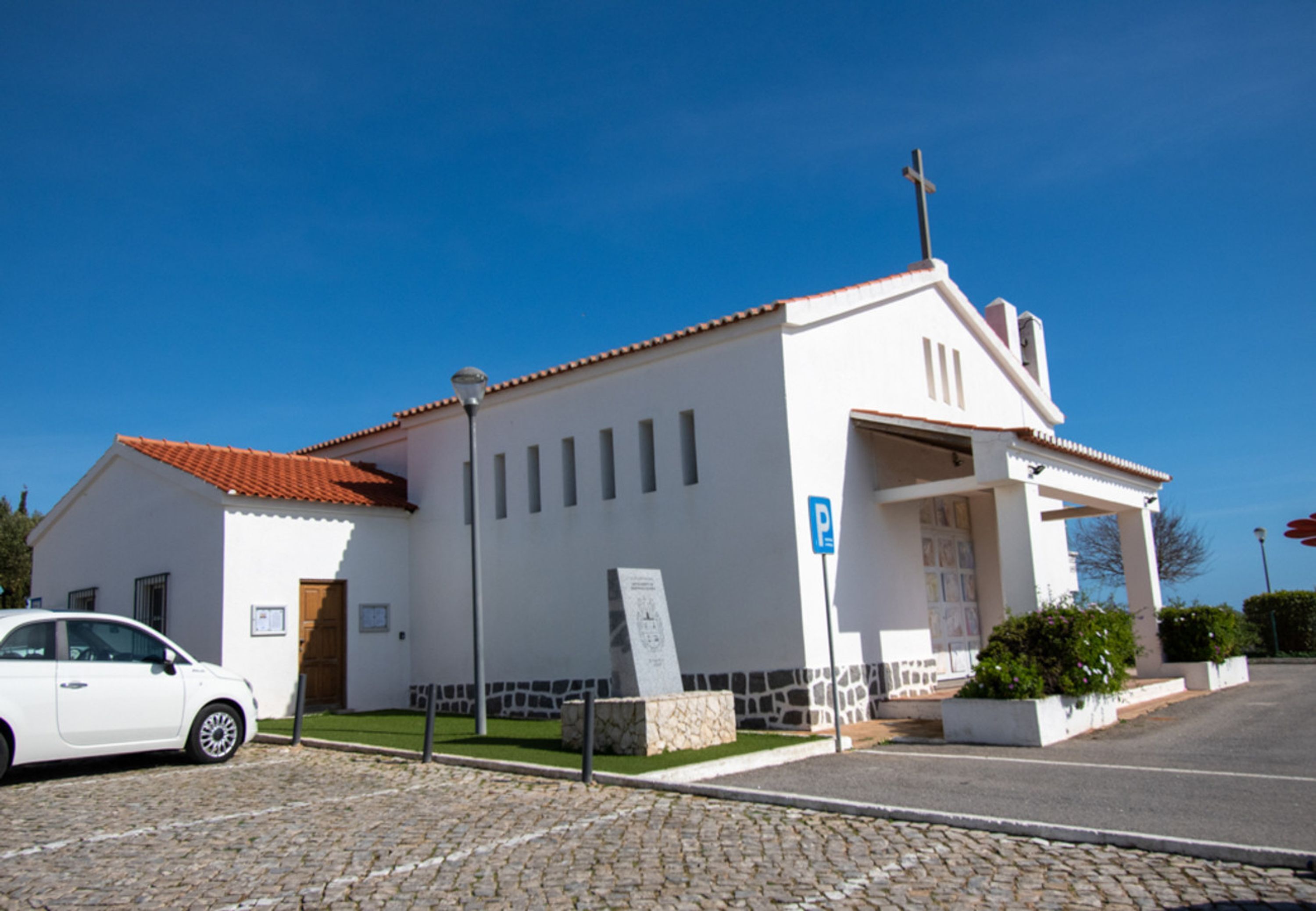 A white church with a red-tiled roof, a parking sign, and a cobblestone street.