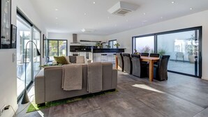 A modern living room with a sofa, dining table, and chairs. A kitchen with a stove and cabinets is visible in the background. - Casa Blanca Magnificent 4 Bedroom Villa With MOD (Lagoa)