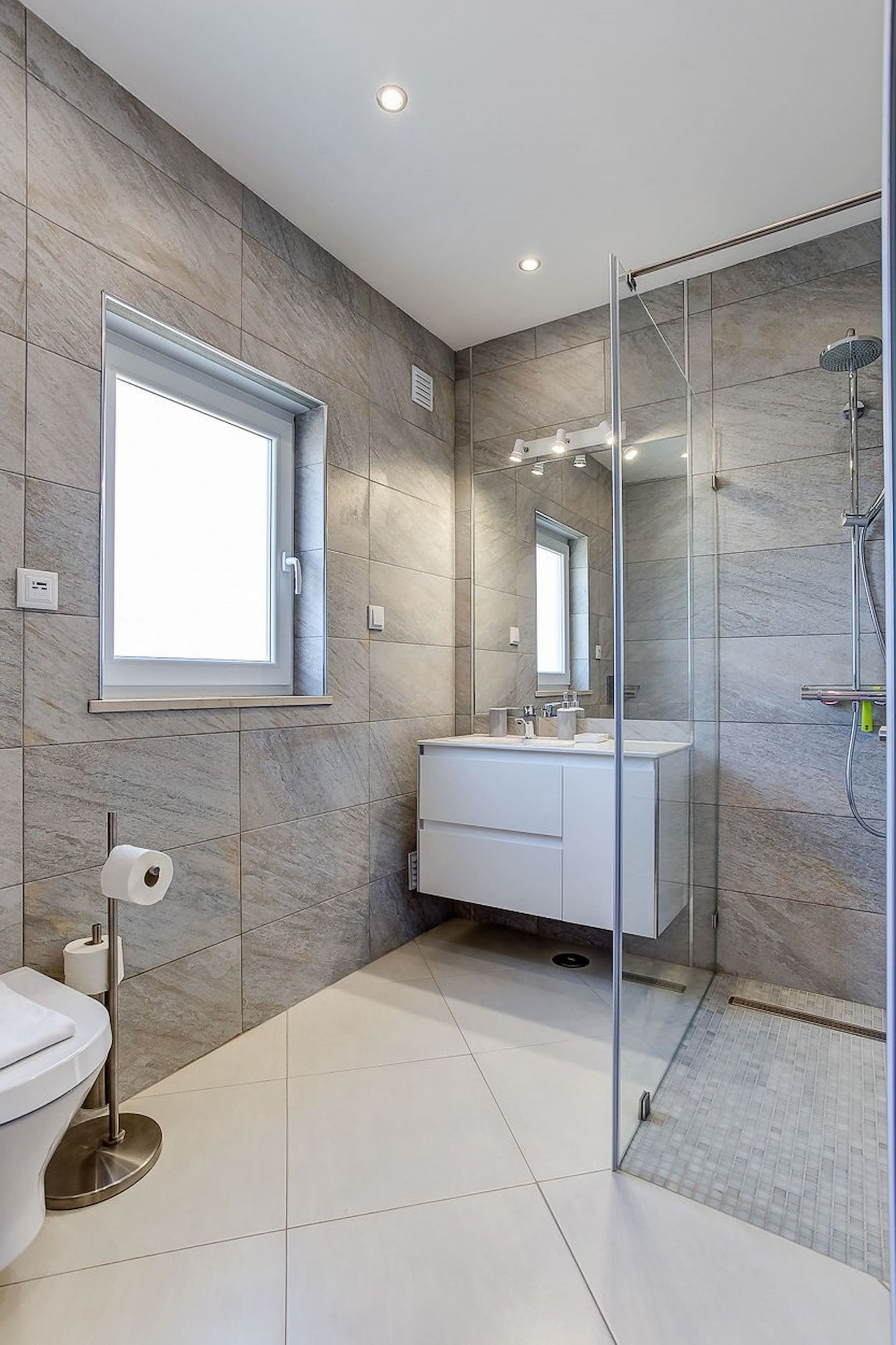 Une salle de bain moderne dotée d’une cabine de douche en verre, d’un grand miroir et d’une fenêtre laissant entrer la lumière naturelle.