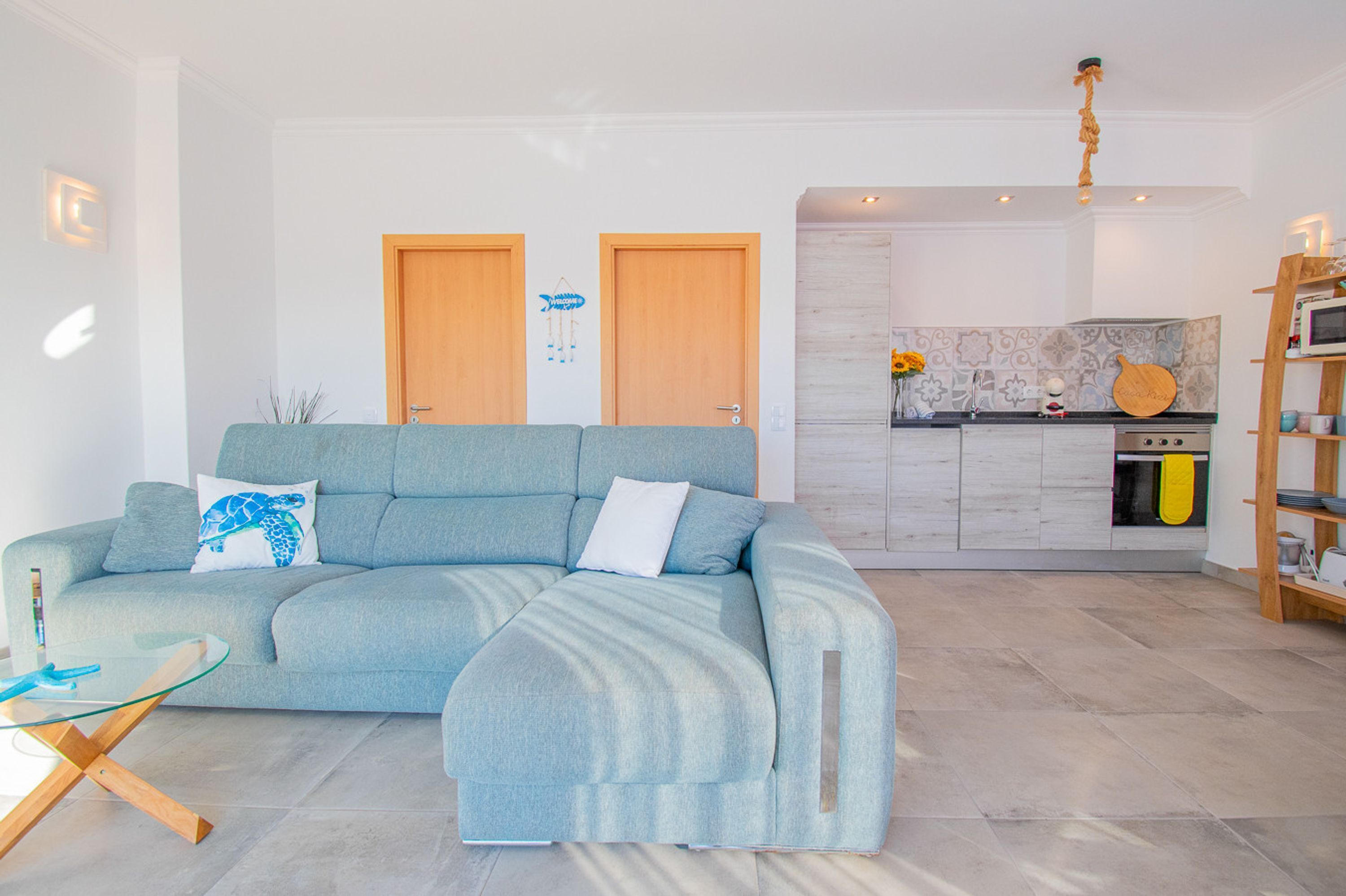 A modern living room with a light blue sofa, a glass coffee table, and a wooden side table. There is a kitchen area with a microwave and a wooden shelf.