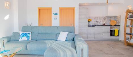 A modern living room with a light blue sofa, a glass coffee table, and a wooden side table. There is a kitchen area with a microwave and a wooden shelf.