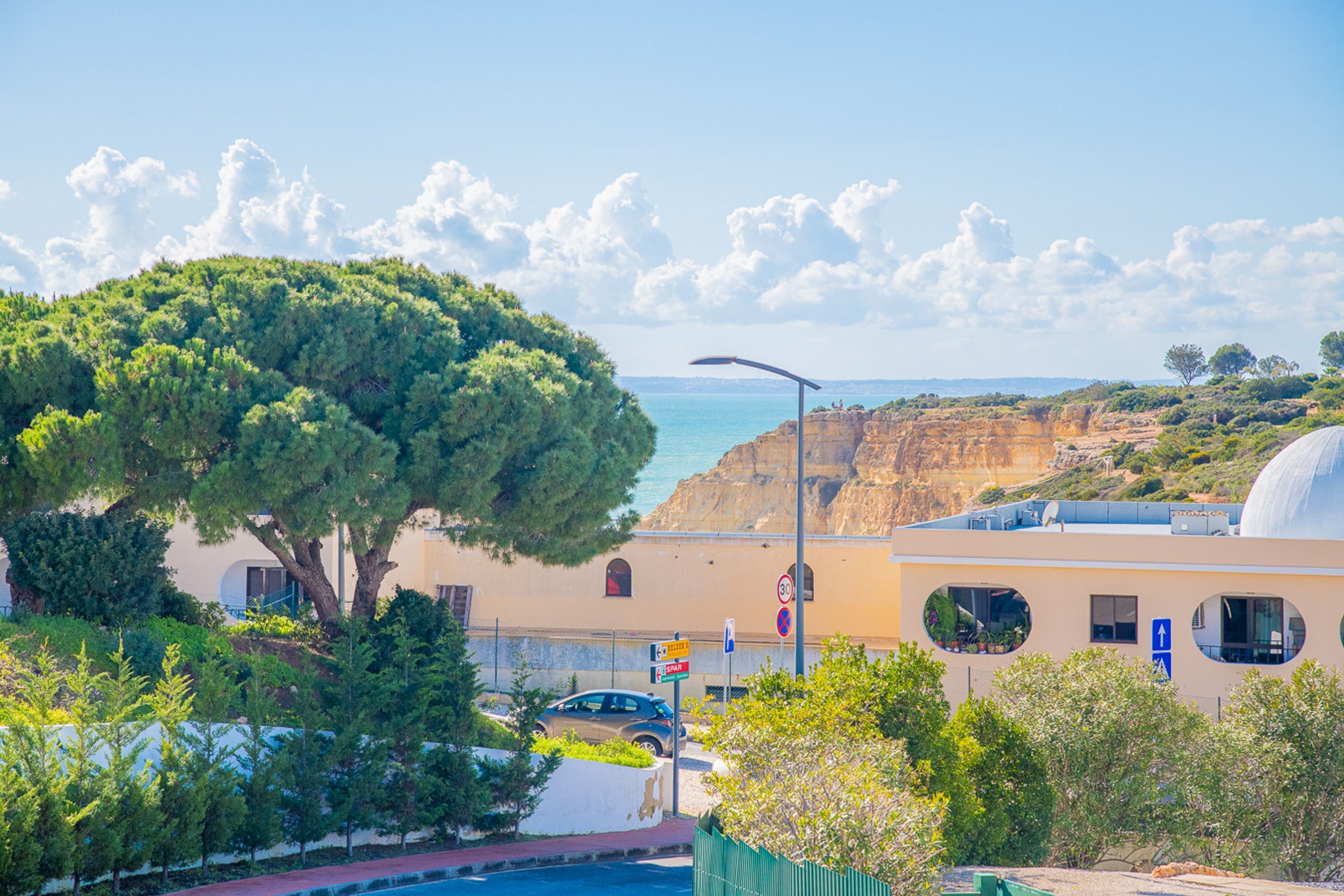 A coastal building with a large tree in front, a car parked nearby, and a clear view of the sea.