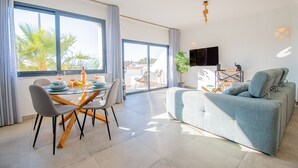 Àmplia zona d'estar amb un sofà gran, taula de dinar amb cadires i televisió penjada a la paret.