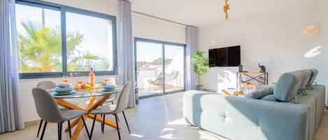 A spacious living area with a large sofa, dining table with chairs, and a TV mounted on the wall.