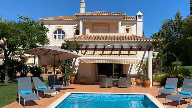 Piscina com espreguiçadeiras, pérgola com toldo listrado e uma casa grande com telhado de cerâmica.