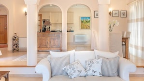 Uma sala de estar espaçosa com um sofá branco, uma mesa de centro de madeira e uma cozinha ao fundo.