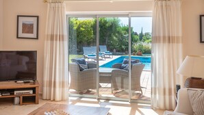 Sala de estar com televisão, mesa de centro e sofá. Porta de vidro deslizante dá acesso a uma área de piscina com espreguiçadeiras.