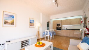 Un salon moderne avec un canapé blanc, une table à manger sur laquelle se trouve un saladier de fruits, et un coin cuisine doté de meubles en bois et d’un réfrigérateur blanc.