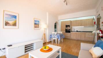 A modern living room with a white sofa, a dining table with a bowl of fruit, and a kitchen area with wooden cabinets and a white refrigerator.
