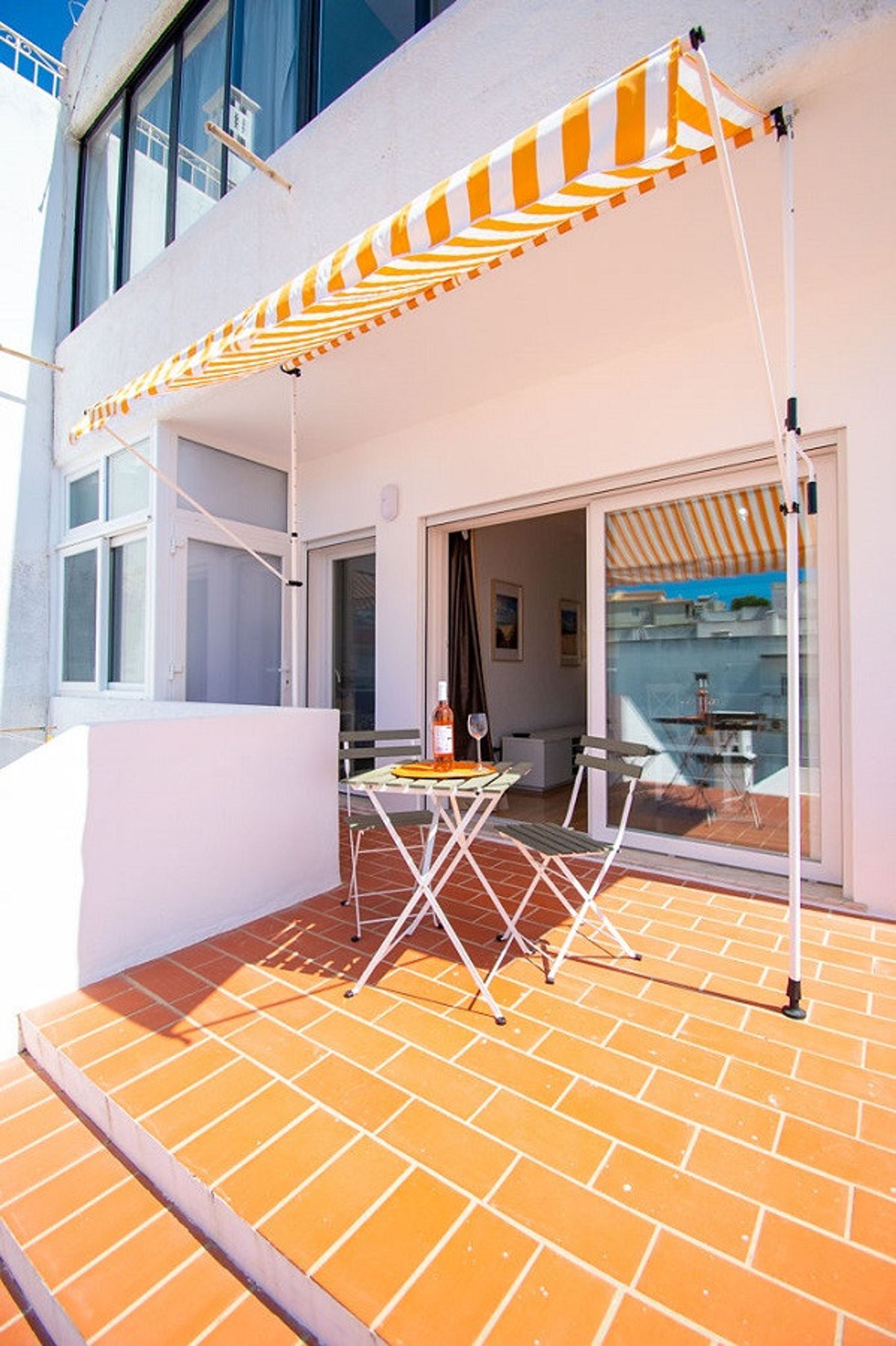 Eine Terrasse im Freien mit gestreiftem Sonnensegel, einem Tisch mit Stühlen und einer Glastür zu einer Küche.