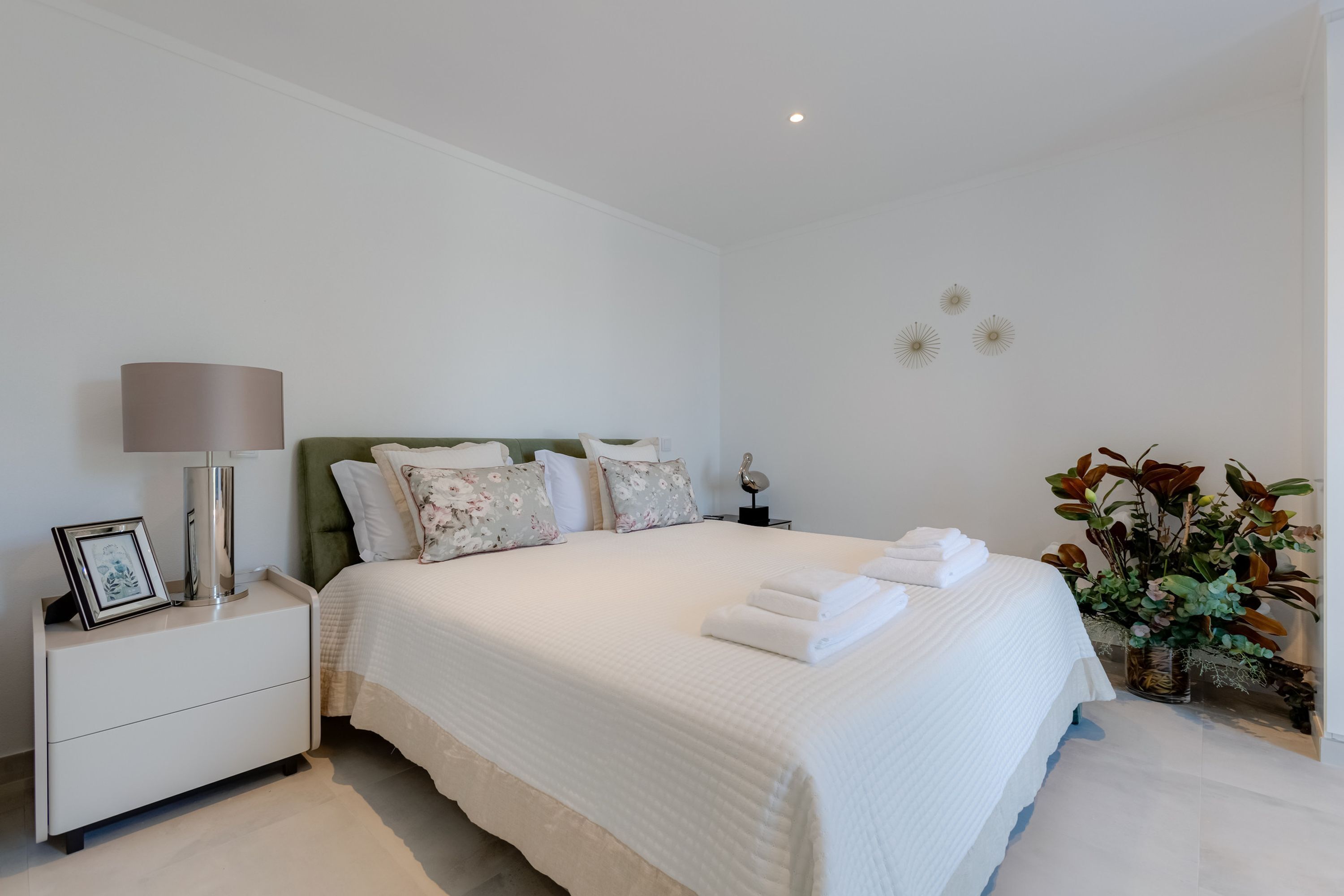 Un lit bien fait, avec du linge de lit blanc et des coussins décoratifs, une lampe de chevet et une table de chevet sur laquelle est posée une photo encadrée.
