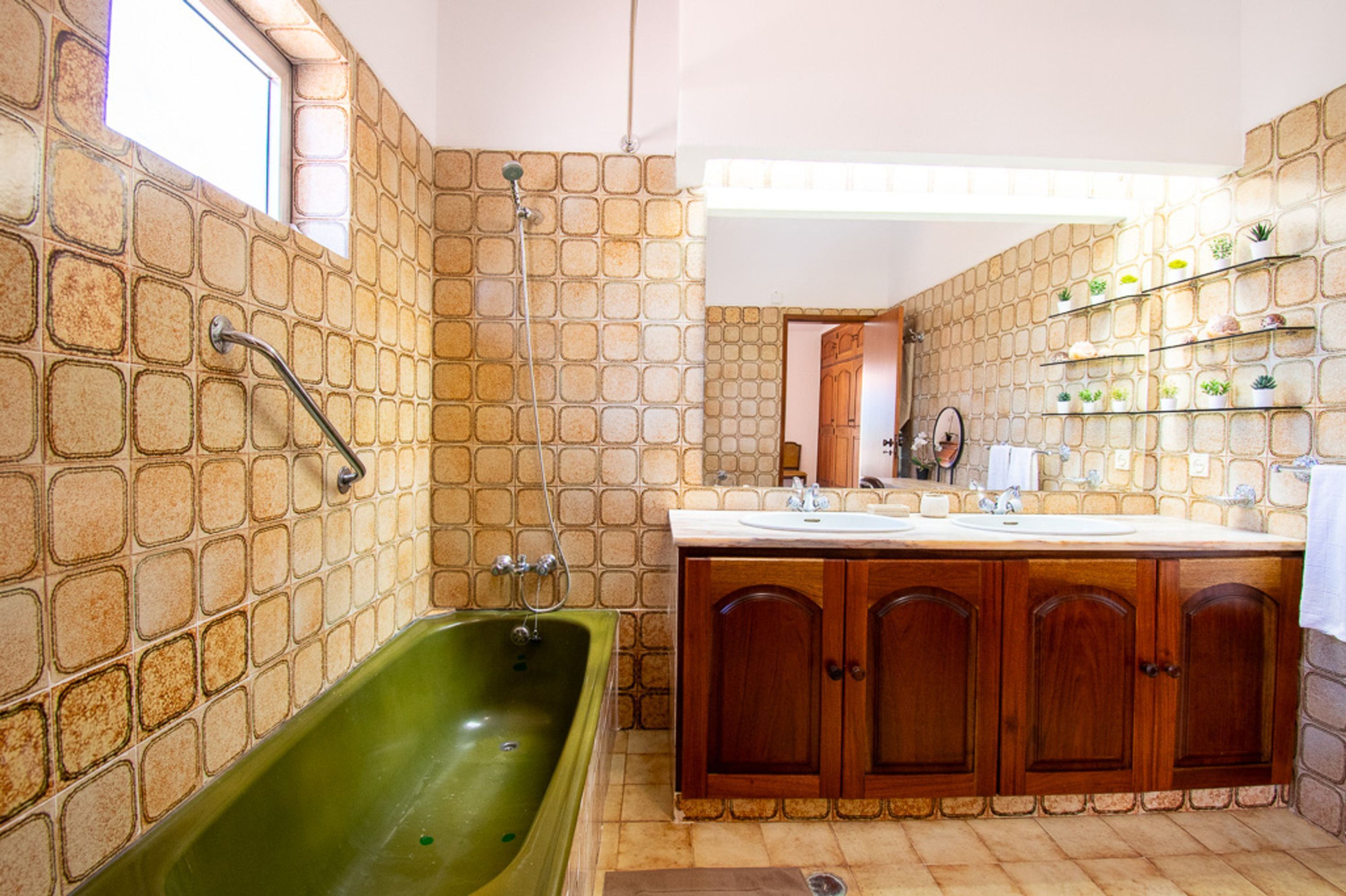 A bathroom with a green bathtub, wooden cabinets, a white sink, and a tiled wall with a window.