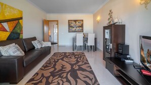A modern living room with a brown sofa, a patterned rug, a dining area with a white table and chairs, and a TV stand with electronic devices. - Lily Fabulous 1 BED APT With Roof Terrace Pool (Lagoa)