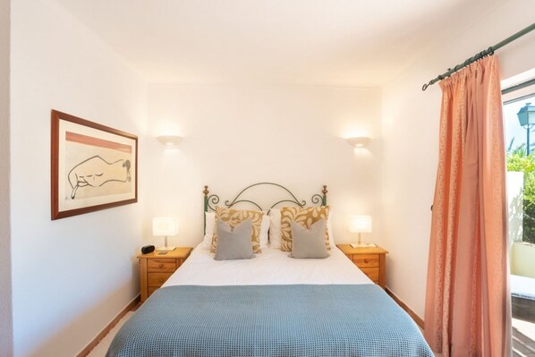 A bedroom with a bed, two bedside tables, a nightstand, a framed picture on the wall, and a window with curtains.