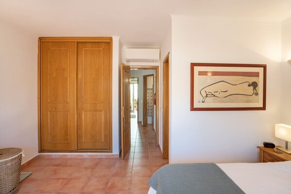 Habitación de hotel con un armario grande de madera, una obra de arte enmarcada en la pared, una mesita de noche con lámpara y una cama con manta gris.