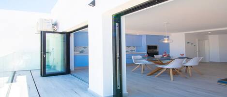 Un interior moderno con zona de comedor, puerta de vidrio que da a un balcón y cocina visible a través del vidrio.