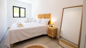 A modern bedroom with a large bed, bedside lamps, a mirror, and a window with a view of trees. - Beach House Apartment (Lagoa)