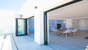 A modern interior with a dining area, a glass door leading to a balcony, and a kitchen visible through the glass. - Beach House Apartment (Lagoa)