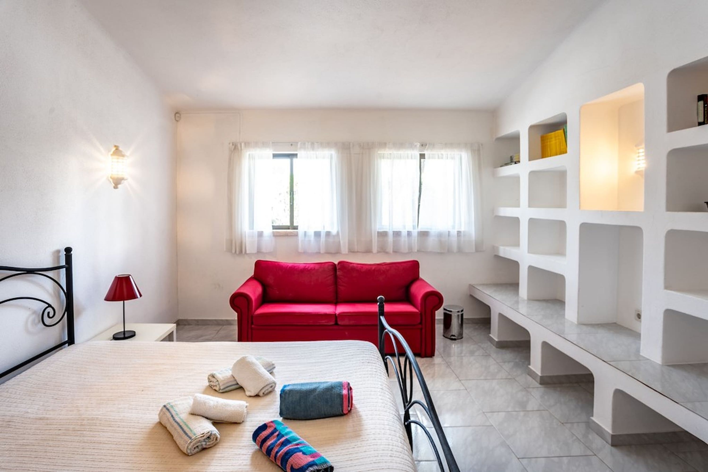 Une chambre d’hôtel avec un lit, un canapé rouge, un mur blanc avec des étagères et une fenêtre avec des rideaux.