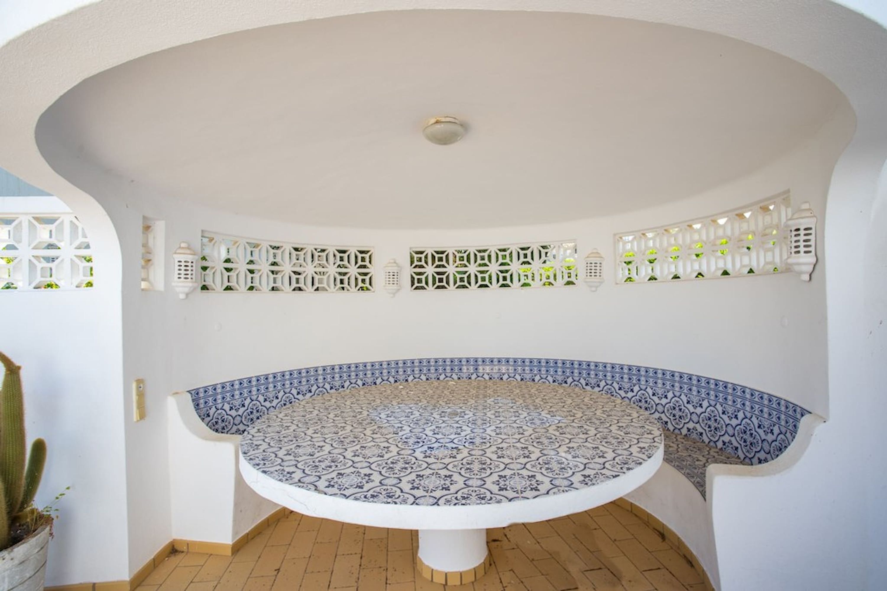 A circular tiled table with a white pedestal base, surrounded by a white wall with decorative lattice patterns and hanging lanterns.