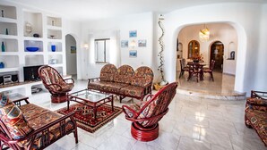 Amplia sala de estar con un sofá, dos sillones y una mesa de centro. Se observa un televisor sobre un mueble bajo, una estantería y un comedor al fondo.