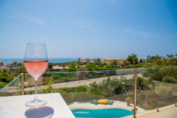 A glass of pink drink on a balcony overlooking a swimming pool and a coastal landscape.