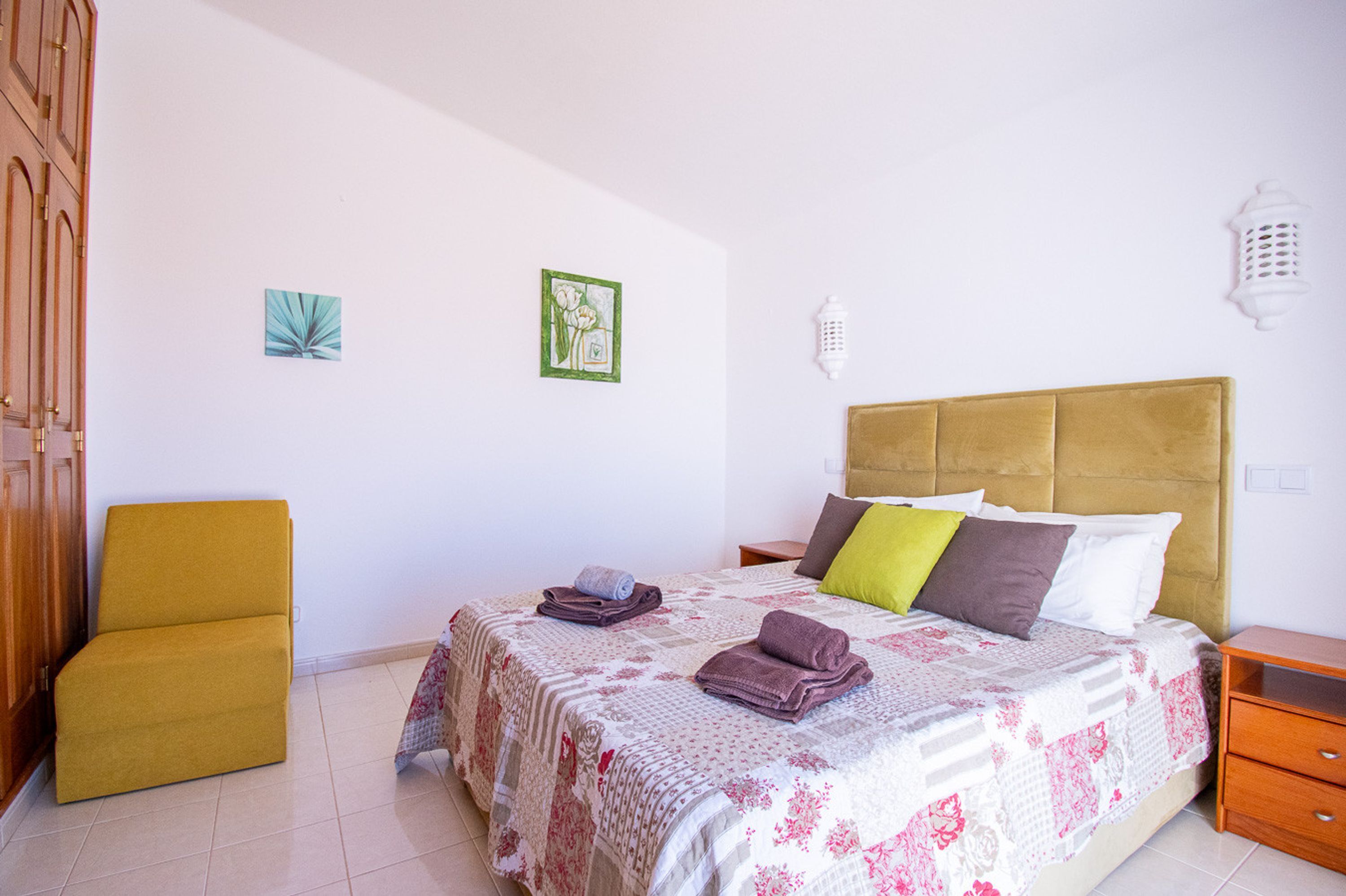 A bedroom with a bed, a chair, a nightstand, and two wall-mounted light fixtures.
