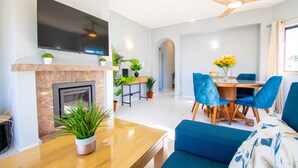 A modern living room with a flat-screen TV, a fireplace, a dining area with blue chairs, and a ceiling fan. - 99B Hibiscus Monte Dourado (Lagoa)
