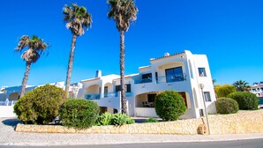 A white building with balconies, surrounded by palm trees and greenery. - 99B Hibiscus Monte Dourado (Lagoa)
