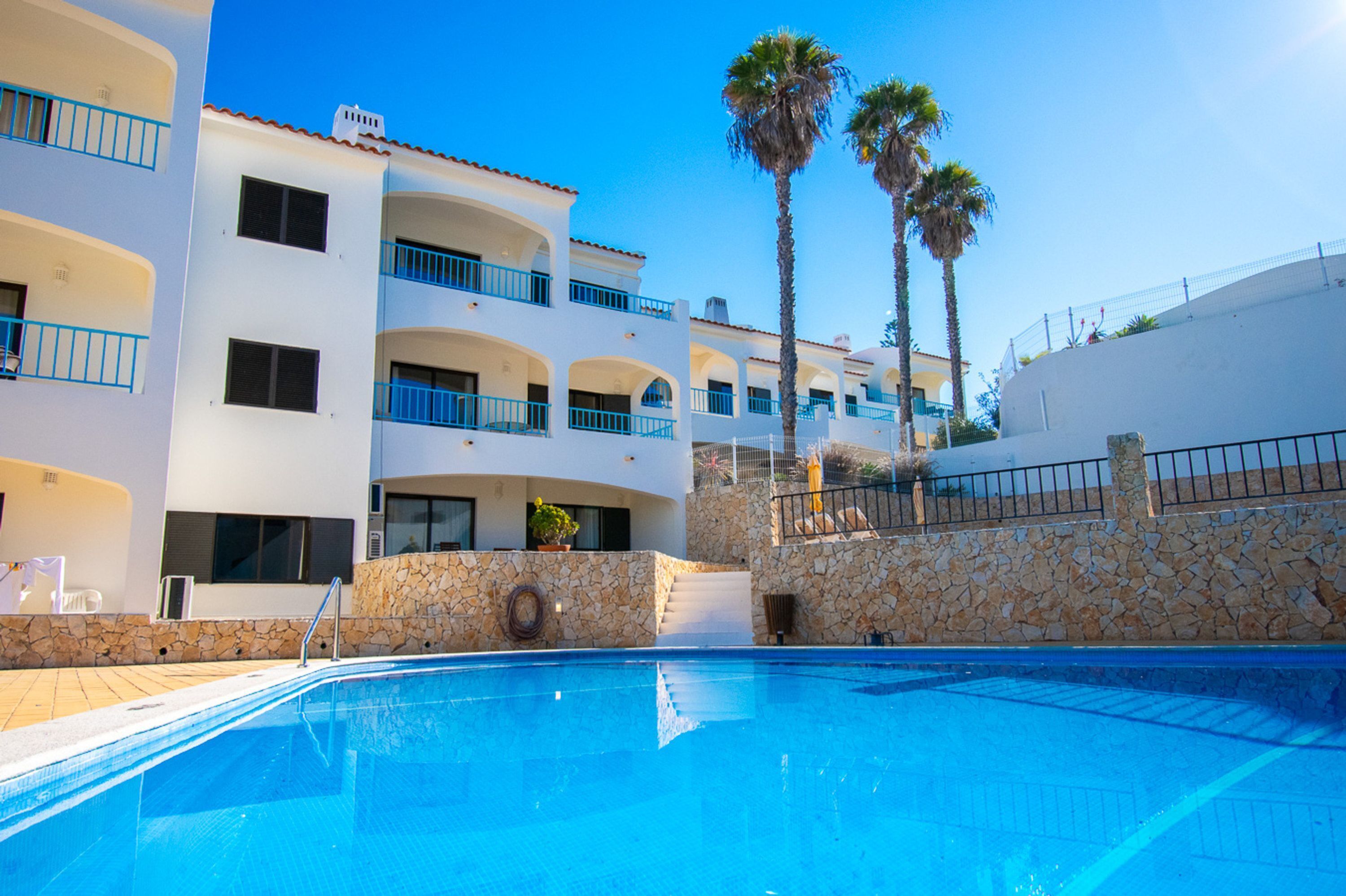 A pool area with a clear blue pool, stone walls, and a white building with balconies and palm trees.