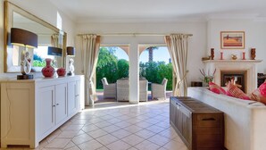 A spacious living room with a white sofa, a coffee table, and a fireplace. There is a large window with curtains, a dining area with chairs, and a cabinet with decorative items. - Casa Marta Three Bedroom Villa With sea Views (Lagoa)