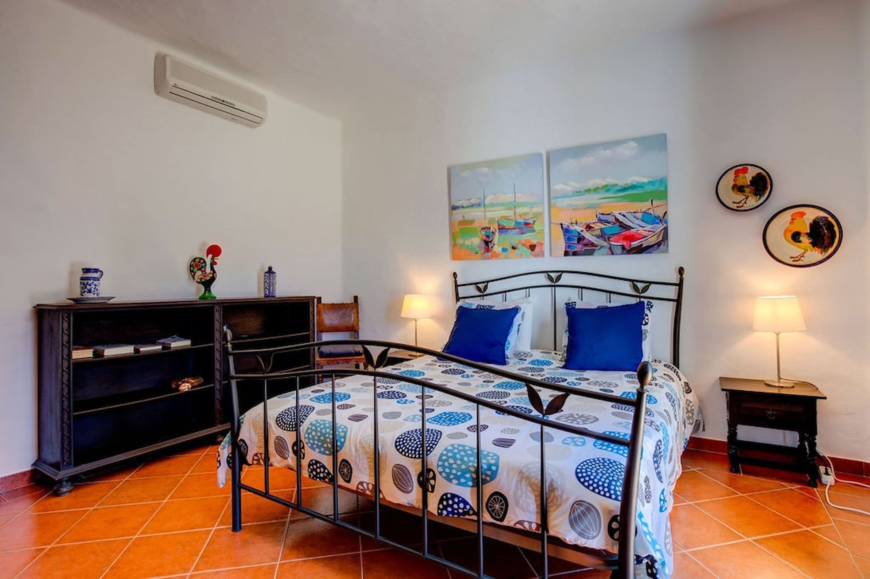 A bedroom with a metal bed frame, a nightstand with a lamp, a dresser, and two wall-mounted paintings.