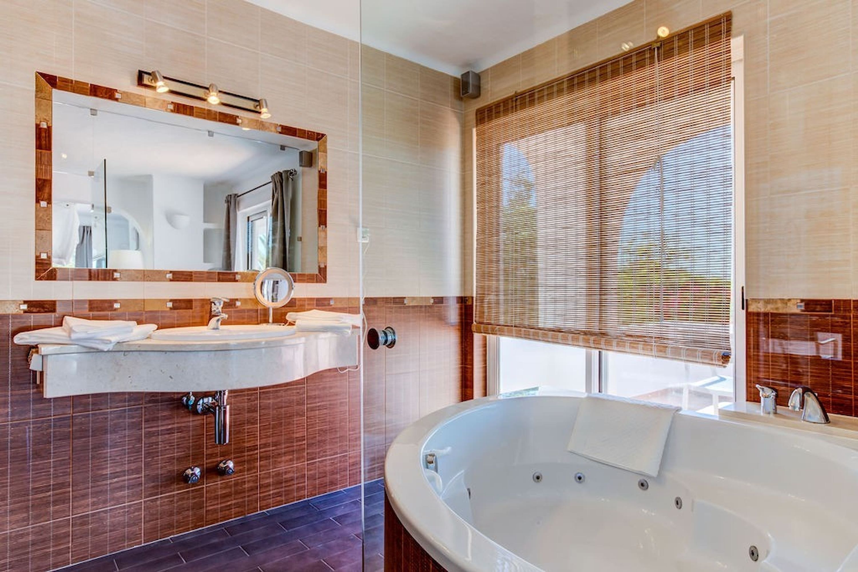 A modern bathroom with a large mirror, two sinks, a bathtub, and a window with blinds.