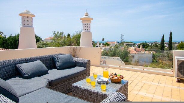 A rooftop terrace with a wicker sofa, a table with drinks, and a view of the sea and buildings. - 67 adm 3 Bedroom Villa Walking Distance to Carvo (Lagoa)