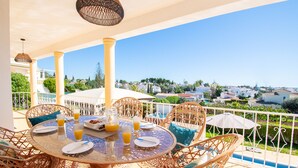 A round table with wicker chairs and a glass top, set on a balcony with a view of a town and greenery.