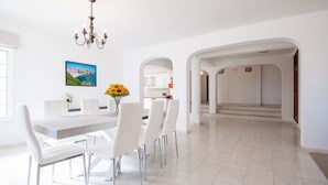 A modern dining area with a white table and chairs, a chandelier, and a view of the kitchen through an archway.