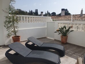 A rooftop terrace with two black wicker lounge chairs, a potted plant, and a white decorative fence.