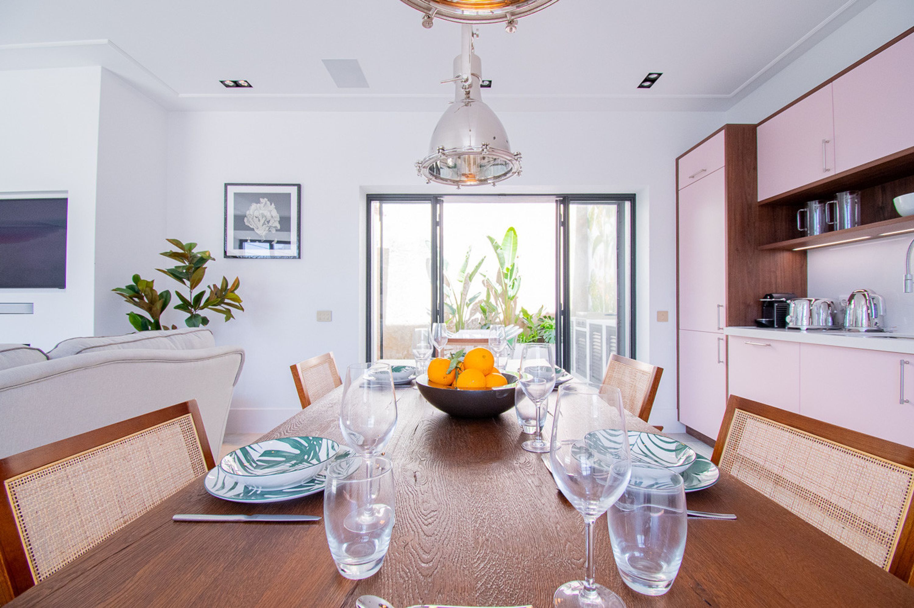 A dining area with a wooden table set for six, surrounded by wicker chairs. A bowl of fruit and wine glasses are on the table.