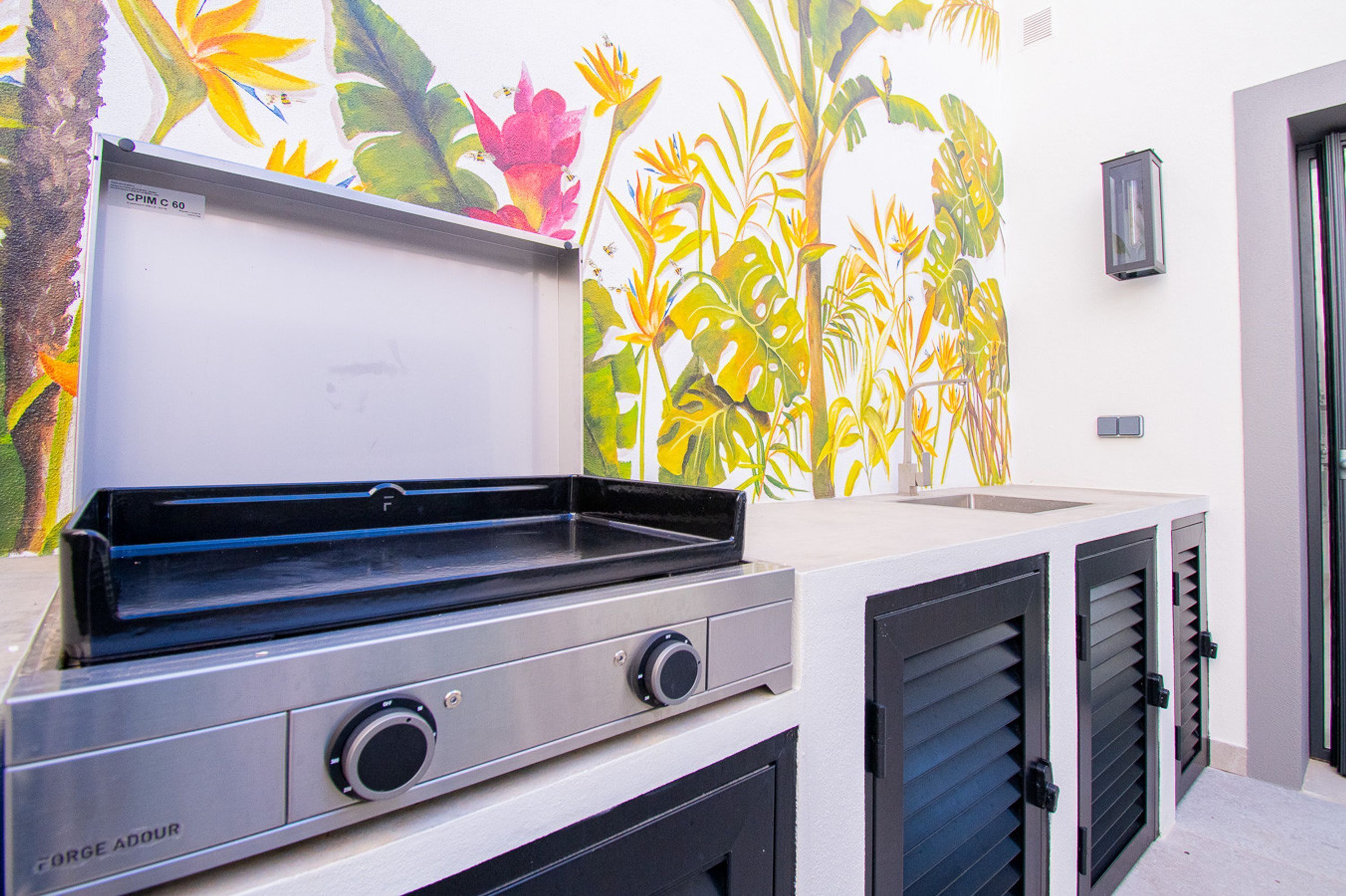 An outdoor kitchen with a grill, a mural of tropical plants, and a wall-mounted light fixture.