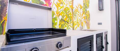 An outdoor kitchen with a grill, a mural of tropical plants, and a wall-mounted light fixture.