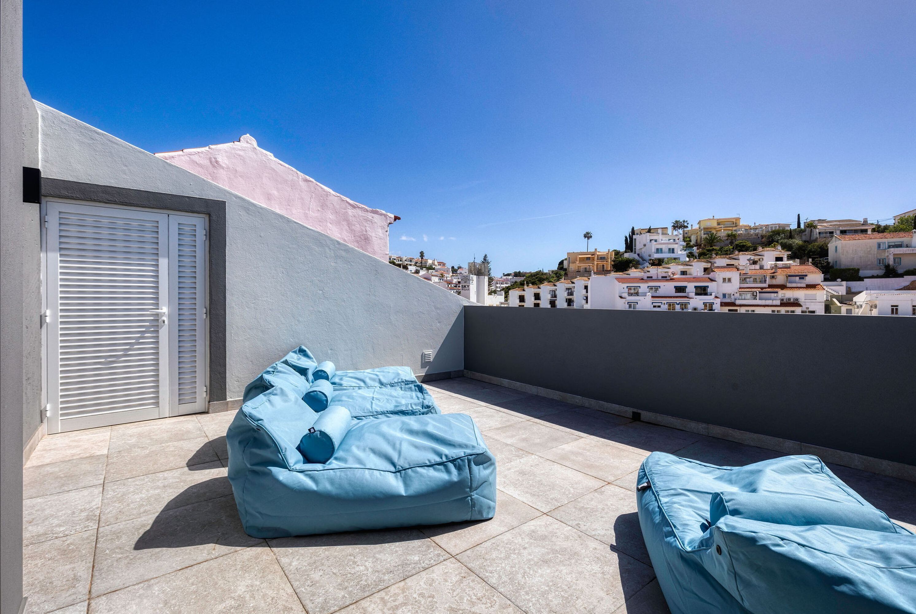 A rooftop terrace with a blue bean bag chair and a covered sofa, a white door, and a view of a coastal town.